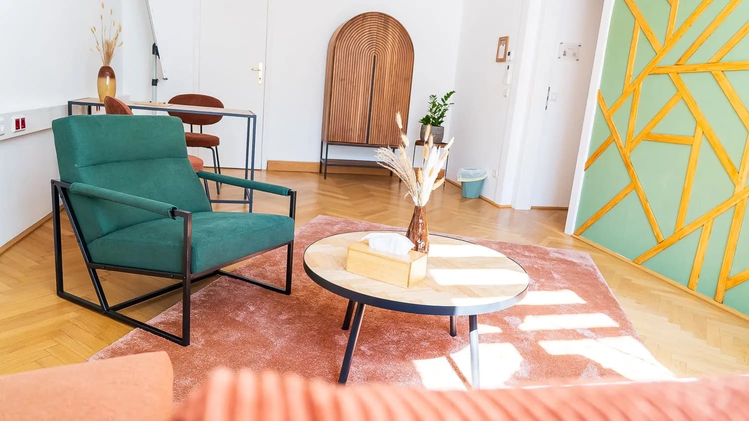 Grüner Sessel und runder Tisch auf einem rosa Teppich, dahinter ein Holzschrank und eine grüne Wand mit Holzleisten.